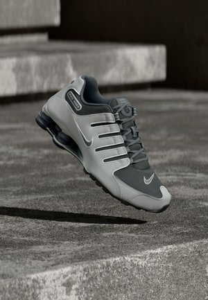 Szaro-czarny sportowy but Nike Shox unoszący się nad teksturowaną betonową powierzchnią z cieniami pod spodem.