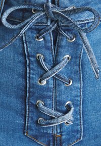 Detalle de prenda de mezclilla que presenta tejido azul, ojales plateados y un diseño de cordones entrecruzados con cordones de mezclilla y costuras en contraste.