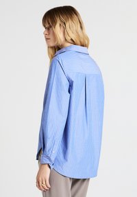 Donna con capelli biondi che indossa una camicia a righe blu e bianca di taglia ampia e pantaloni beige, rivolta a sinistra su uno sfondo neutro.