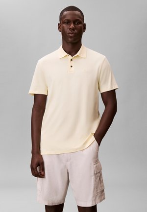 Man draagt een lichtgeel poloshirt met korte mouwen en beige cargoshorts, staand tegen een effen grijze achtergrond.