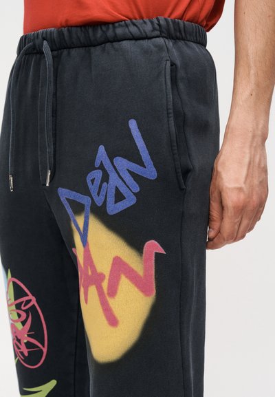 Κοντινή λήψη μαύρων αθλητικών παντελονιών με πολύχρωμα γράμματα και σχέδια σε στυλ γκράφιτι κοντά στην δεξιά τσέπη, φορεμένα με κόκκινο πουκάμισο.