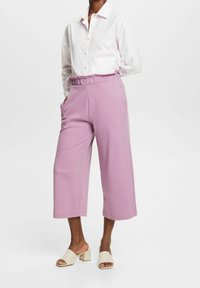 Personne portant une chemise blanche à boutons, un pantalon large lilas taille haute et court, ainsi que des sandales beige à talons blocs et bouts ouverts.