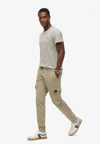 Grijze katoenen T-shirt, khaki cargobroek met zijzakken en witte sneakers met zwarte accenten. Model staat zelfverzekerd met de handen in de zakken.