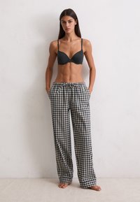 Vrouw die blote voeten tegen een witte muur staat, draagt een zwarte bh en losse geruite pyjamabroek met haar handen in de zakken.