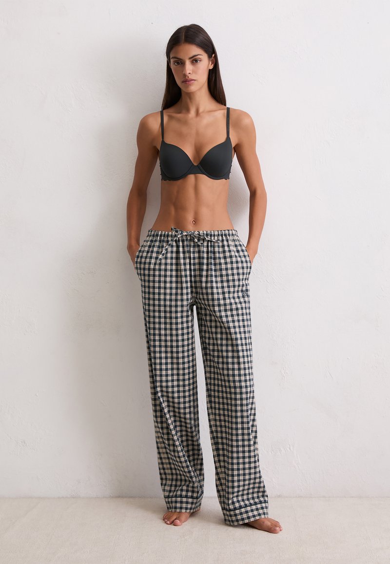 Vrouw die blote voeten tegen een witte muur staat, draagt een zwarte bh en losse geruite pyjamabroek met haar handen in de zakken.