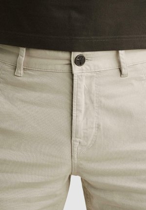 Hellbeige Hose mit sichtbaren Gürtelschlaufen, vorderen Taschen-Nähten und einem Metallknopfverschluss, kombiniert mit einem dunkelbraunen Hemd.