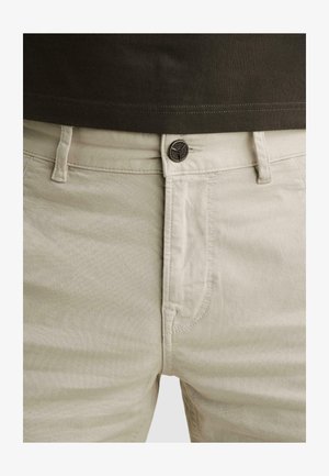 Pantalon beige clair avec passants de ceinture visibles, coutures des poches avant et fermeture par bouton métallique, associé à une chemise marron foncé.
