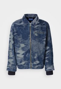 Chaqueta de mezclilla azul con un patrón texturizado, cierre frontal con cremallera, bolsillo en el pecho y puños acanalados. Marca visible en la tela.