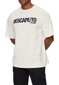 Korte mouwen wit katoenen t-shirt met een opvallende marineblauwe grafische tekst "#ESCAPE/TO PARADISE" op de borst. Ontspannen pasvorm.