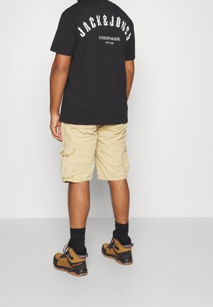 Person iført sort Jack & Jones T-shirt, beige cargoshorts, sorte sokker og brune vandrestøvler, står med ryggen mod kameraet.