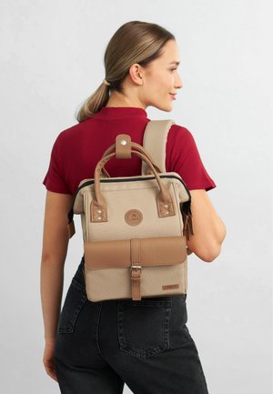 Sac à dos en toile beige avec des accents en cuir marron, de forme rectangulaire, avec des poignées supérieures, des bretelles réglables et un fermoir décoratif.