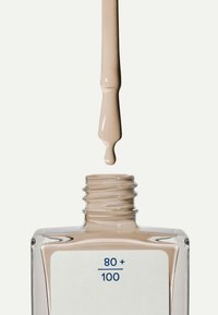 Botella de esmalte de uñas de cristal transparente con forma cuadrada. Un esmalte beige claro gotea de un pincel largo, que presenta una etiqueta blanca con texto azul.