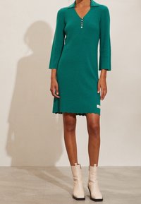 Robe côtelée bleu sarcelle avec un col en V, une patte de boutonnage, des manches trois-quarts et un ourlet festonné. Portée avec des bottines blanches à la cheville.