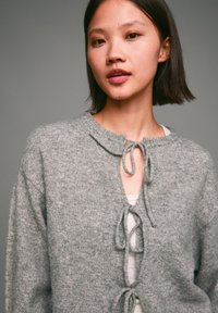 Szary sweter na drutach z okrągłym dekoltem, z wycięciem w kształcie klucza i wiązaniami z przodu. Miękka tekstura i luźny krój.