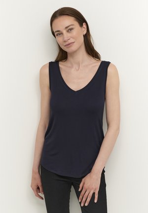Femme aux longs cheveux bruns portant un haut sans manches bleu marine à col en V et un pantalon noir, debout contre un mur blanc.