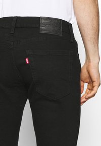 Jeans de mezclilla negra con una etiqueta de cuero en la cinturilla y una etiqueta roja. El bolsillo trasero tiene un patrón de costura distintivo. Textura suave.