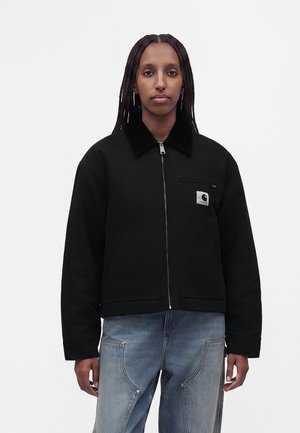 Personne avec de longues tresses portant une veste noire zippée avec une poche poitrine et un logo Carhartt, associée à un jean bleu clair.