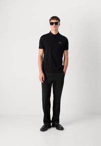 Polo noir à manches courtes avec logo brodé, associé à un pantalon noir et des lunettes de soleil sombres, se tenant sur un fond blanc.