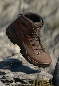 Botas de caminhada castanhas com uma superfície texturizada, material sintético durável, biqueira reforçada e design de atacadores, posicionadas contra um fundo de rochas naturais.