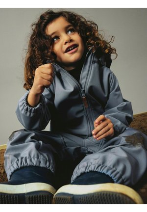 Barn med krøllete hår iført en grå genser med glidelås og marineblå sko, sitter og ser opp med et tankefullt uttrykk.