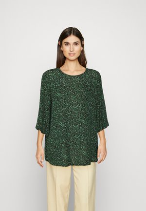Blusa verde com padrão floral, com mangas três quartos, decote redondo e um corte solto, combinada com calças de cor creme.
