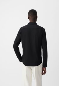 Camisa negra de manga larga con cuello clásico, puños abotonados y una textura suave, combinada con pantalones de color claro.