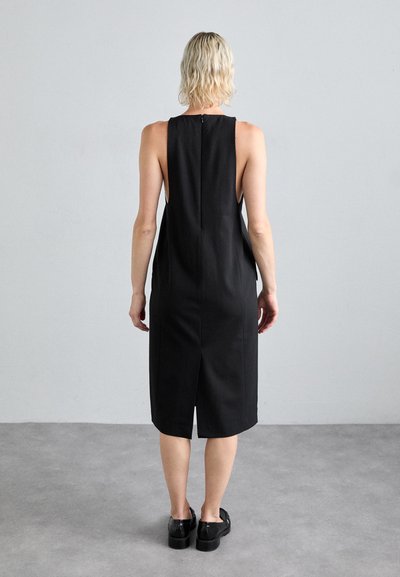 Schwarzes ärmelloses Kleid mit einer taillierten Silhouette, seitlichen Schlitzen und einem Reißverschluss auf der Rückseite. Der Stoff wirkt glatt und matt. Träger mit schwarzen Schuhen.