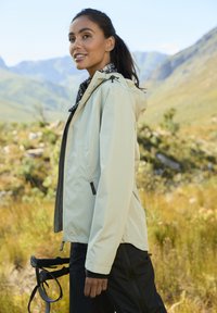 Šviesiai smėlio spalvos atsparus vandeniui džemperis su kapišonu, reguliuojamomis stygomis ir užtrauktukais uždaromomis kišenėmis. Derinama su tamsiomis kelnėmis ir raštuota šaliku.