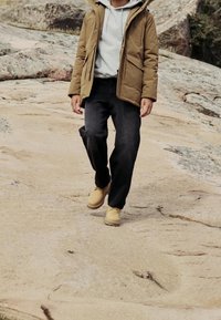 Personne portant une veste beige, un sweat à capuche gris, un pantalon noir et des bottes beige marchant sur un terrain rocheux en plein air.