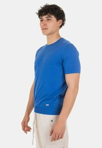 Maglione lavorato a maglia blu a maniche corte, collo rotondo, polsini e orlo a coste, texture liscia, con un piccolo logo in basso a sinistra.