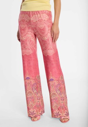 Femme portant un pantalon large rose avec des motifs paisley complexes et des talons ouverts roses, assortis à un haut sans manches jaune.
