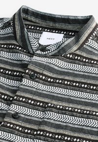 Chemise boutonnée rayée noire, blanche et grise avec des motifs géométriques et un col mandarin, montrant l'étiquette de la marque « NEXT » à l'intérieur du col.