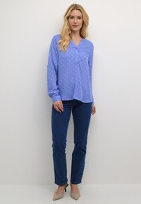 Blouse à manches longues bleu clair avec des pois blancs, décolleté en V et détails de boutons, associée à un jean bleu foncé et des escarpins beiges.