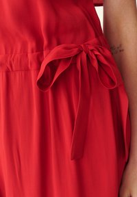 Rød stof jumpsuit med samlet talje og stort bue-detalje på siden. Glat tekstur og let design, der fremhæver den levende farve.
