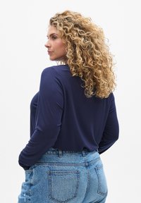 Navy long-sleeve top in soft fabric, relaxed fit. Worn with high-waisted light blue denim jeans featuring back pockets. Textured, wavy hair.