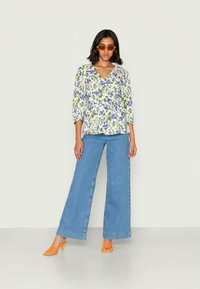 Mulher a usar óculos de sol laranja, blusa floral com flores roxas, calças de ganga largas azuis e sandálias de salto laranja, apoiada numa parede lisa.