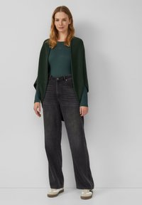 Dunkelgraue weite Jeans mit hoher Taille, kombiniert mit einem dunkelgrünen Strickoberteil und einer übergroßen Strickjacke. Lässiges Outfit mit Sneakers.