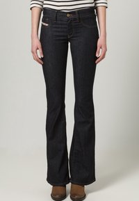 Ciemnoniebieskie dzwony z klasycznym pięciopocketowym wzorem, z zapięciem na guzik i kontrastującymi przeszyciami. Gładka tekstura dżinsu.