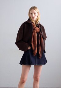 Femme blonde portant une grande veste marron foncé, une écharpe couleur rouille et une jupe en denim court foncé, se tenant contre un mur clair et simple.
