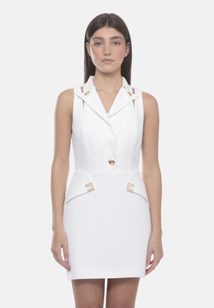 Donna in piedi di fronte, indossa un vestito bianco senza maniche con bottoni dorati e accenti dorati geometrici sulle tasche in vita.