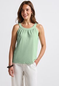 Světle zelený bezramenný top s kulatým výstřihem a nařasením, kombinovaný s bílými kalhotami. Hladká textura.