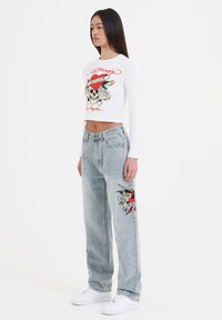 Hvid langærmet cropped skjorte med grafisk print af et kranium, parret med lyseblå baggy jeans med farverige broderede lapper.