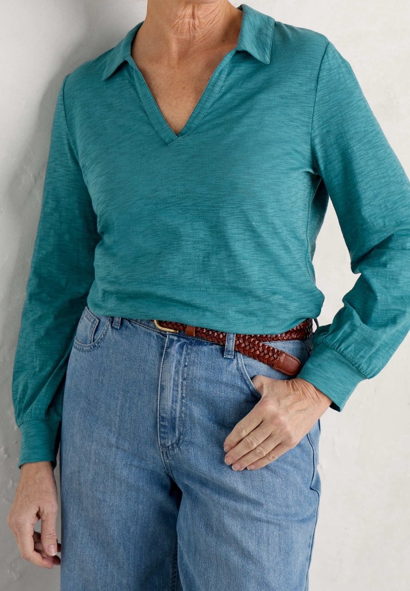 Blusa lunga teal a maniche lunghe con scollatura a V e tessuto testurizzato, abbinata a jeans blu a vita alta e una cintura in pelle intrecciata marrone.