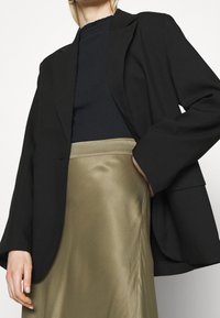 Blazer structuré noir porté sur un haut côtelé bleu marine, associé à une jupe en soie vert olive. Caractéristiques : manches longues et coupe décontractée.