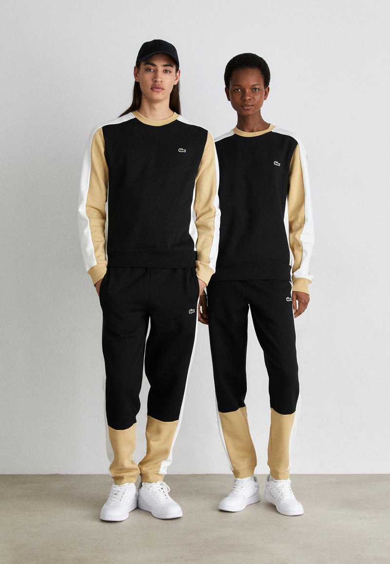 Ensembles de sport noirs et beiges comprenant des manches longues, des panneaux contrastants, un col rond et des accents de logo, associés à des baskets blanches.