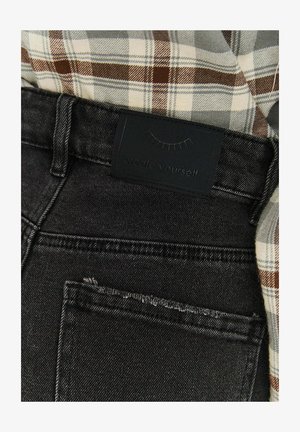 Schwarze Denim-Jeans mit strukturierter Oberfläche, ausgestattet mit einem dunklen Markenlabel am Bund, teilweise bedeckt von einem braunen karierten Hemd.