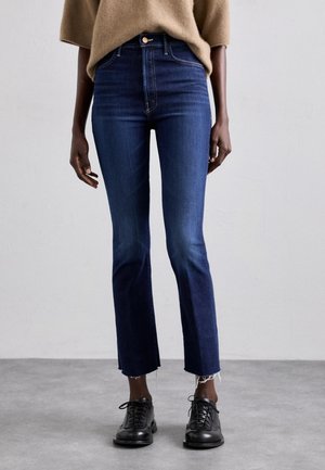 Person iført mørkeblå højttaljede jeans med rå kanter og sorte snøresko, stående mod en ensfarvet grå baggrund.