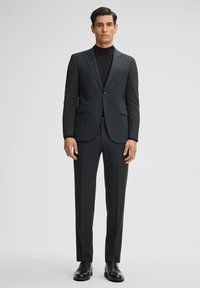 Donkergrijs op maat gemaakt pak met een notch lapel, een enkelgeknoopte jas en slim-fit broek. Gedragen over een zwarte turtleneck, aangevuld met zwarte schoenen.