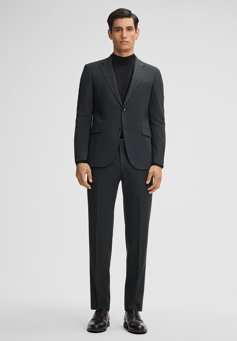 Donkergrijs op maat gemaakt pak met een notch lapel, een enkelgeknoopte jas en slim-fit broek. Gedragen over een zwarte turtleneck, aangevuld met zwarte schoenen.