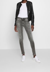 Persona con una chaqueta de cuero negra, camiseta blanca, jeans ajustados grises desgastados y zapatillas blancas de pie contra un fondo gris.